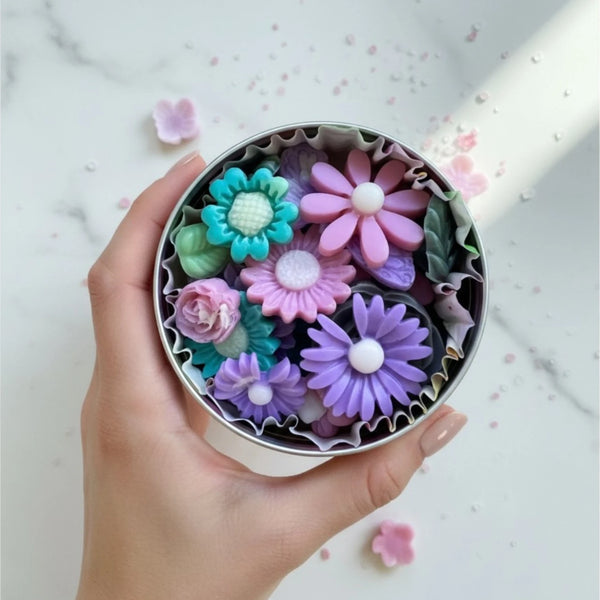 A hand holds a round metal tin filled with colorful flower-shaped soap bars in pastel shades of purple, pink, teal, and green, set against a light marble background with scattered flower petals.