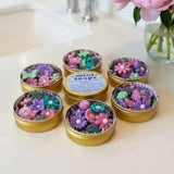 Six small round gold tins filled with colorful flower-shaped mini soaps, arranged on a light countertop near a sink with flowers in the background. One tin has a visible label reading 'mini soaps'.