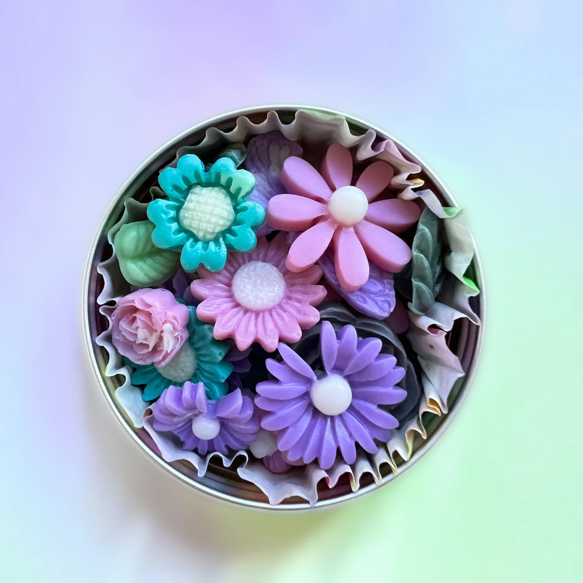 A top-down view of a round metal tin filled with colorful flower-shaped soaps, set against a soft pastel gradient background.