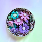 A top-down view of a round metal tin filled with colorful flower-shaped soaps, set against a soft pastel gradient background.