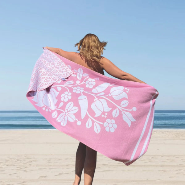 Woman standing on the beach with her back to the camera, holding a pink towel stretched out behind her.