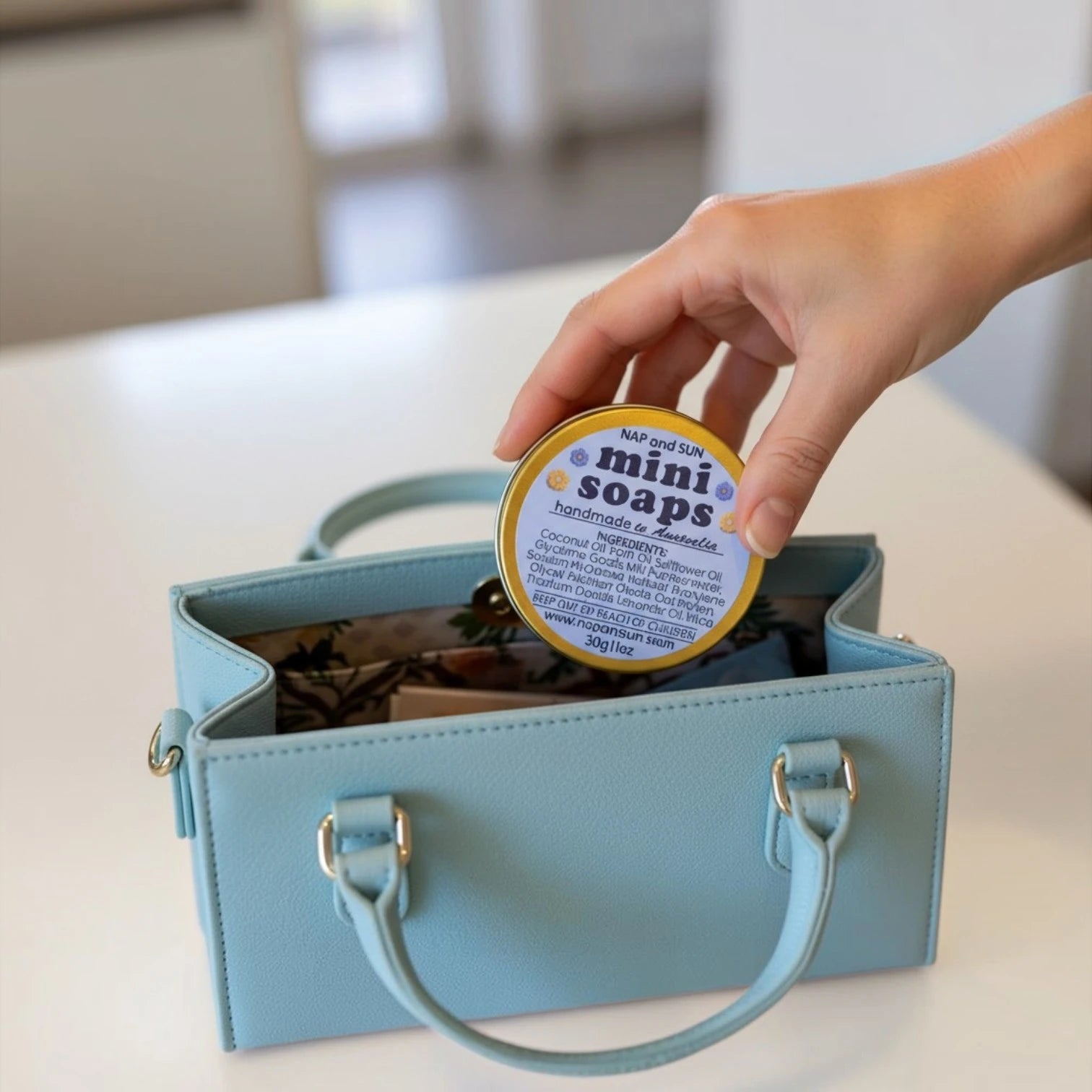 A hand is holding a golden tin with text ' mini soaps' and putting it into a light blue handbag.