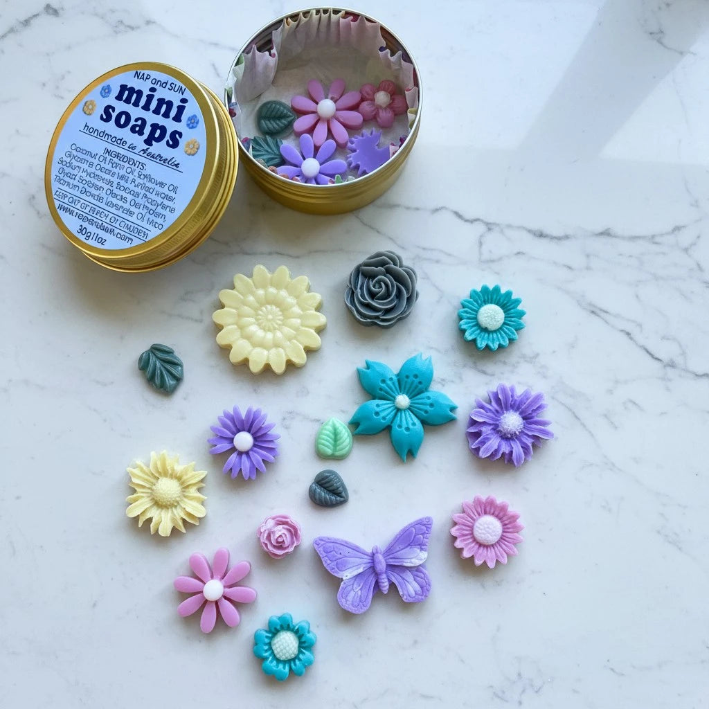 A round gold tin labeled 'mini soaps' with several colorful flower and butterfly-shaped handmade soaps arranged on a white marble surface. The tin is open, showing more soap pieces inside.