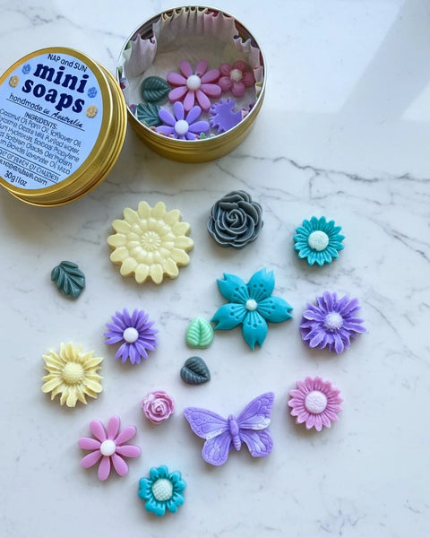 A round gold tin labeled 'mini soaps' with several colorful flower and butterfly-shaped handmade soaps arranged on a white marble surface. The tin is open, showing more soap pieces inside.