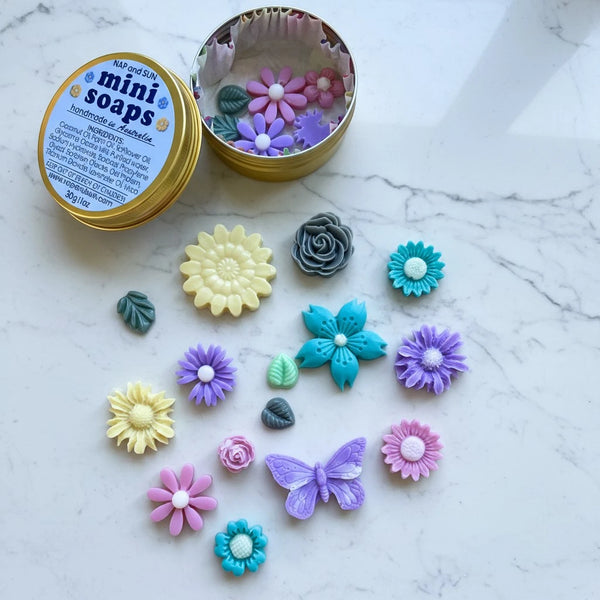 A round gold tin labeled 'mini soaps' with several colorful flower and butterfly-shaped handmade soaps arranged on a white marble surface. The tin is open, showing more soap pieces inside.