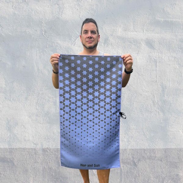 Man standing in front of a wall, facing the camera, holding an indigo-coloured gym towel with a black star pattern, stretched out with both hands.