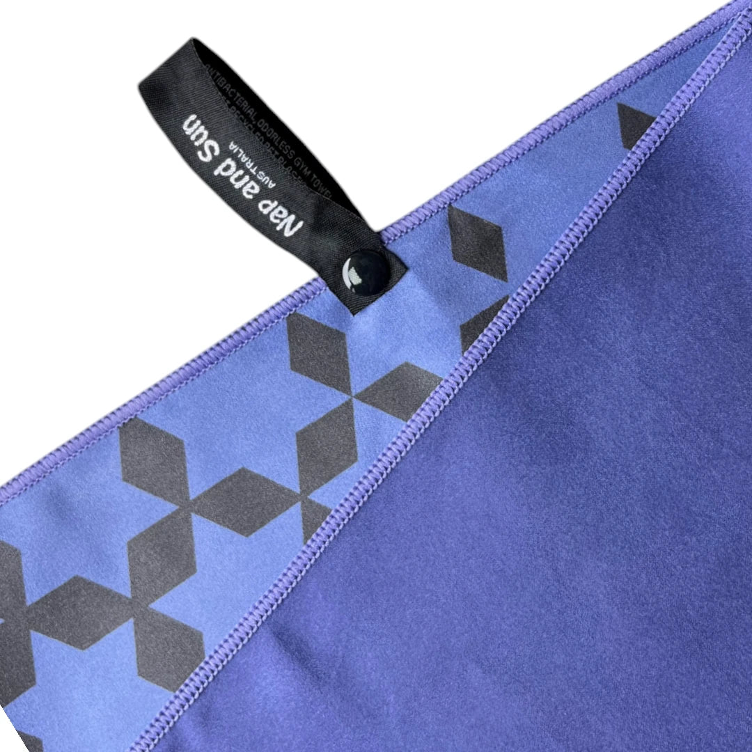Close-up of Nap and Sun indigo blue gym towel showing the front and back. The front is a light blue with a black star pattern, while the back is a darker indigo blue. A black hook with a press stud is attached for easy hanging.