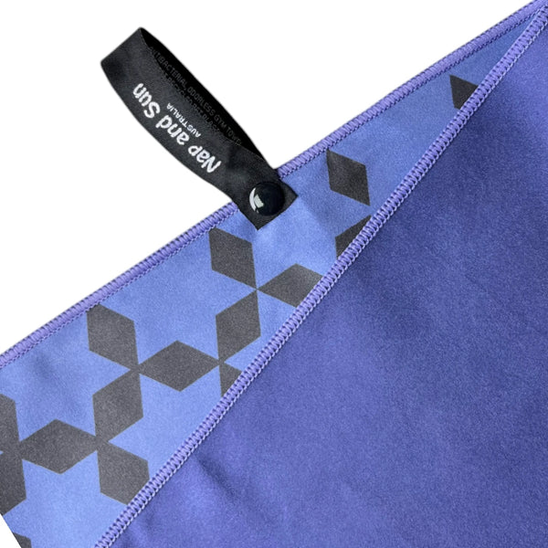 Close-up of Nap and Sun indigo blue gym towel showing the front and back. The front is a light blue with a black star pattern, while the back is a darker indigo blue. A black hook with a press stud is attached for easy hanging.