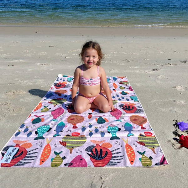 Child sitting on a colorful beach towel with a sea-themed pattern on a sandy beach.