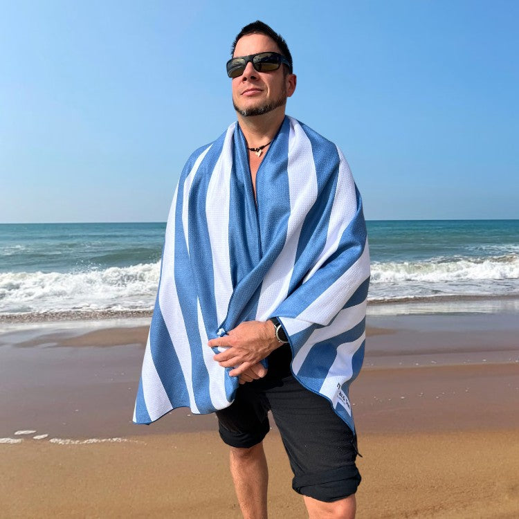 Man standing on a beach next to the sea, wrapped in a blue and white striped beach towel. Nap and Sun Stripe Love towel, free wetbag included