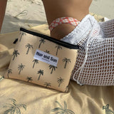 Close-up of the Sunset Palms mustard yellow beach towel with palm tree patterns, laid out on the sand. A woman is sitting on the towel, with the matching wetbag resting against her side.