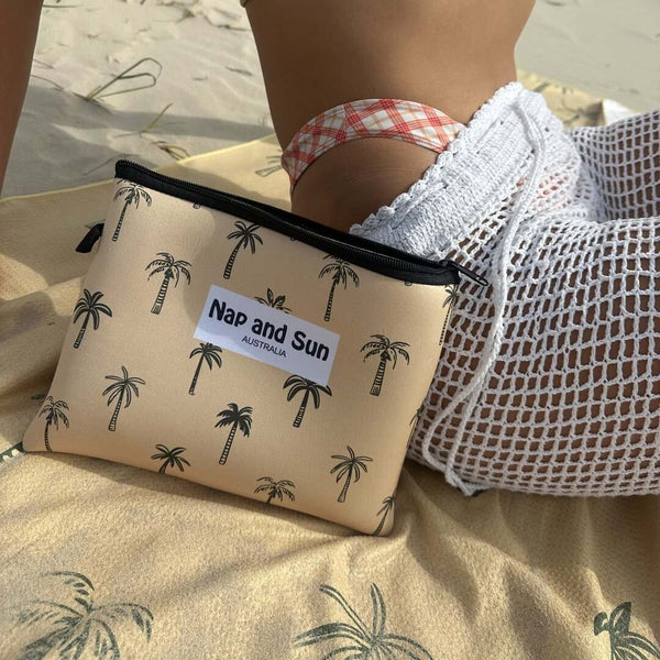 Close-up of the Sunset Palms mustard yellow beach towel with palm tree patterns, laid out on the sand. A woman is sitting on the towel, with the matching wetbag resting against her side.