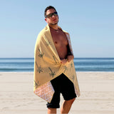 Man stands on soft sand next to the sea, facing the camera. He is wrapped in a yellow-gold palm tree pattern Nap and Sun Sunset Palm beach towel.
