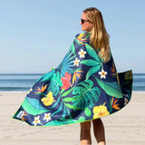 Woman standing on the beach with her back to the camera, wrapped in a Tropical Bliss towel featuring green tropical leaves and hibiscus flowers.