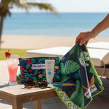 Tropical-themed towel with pouch, and drink on a table with a beach background.
