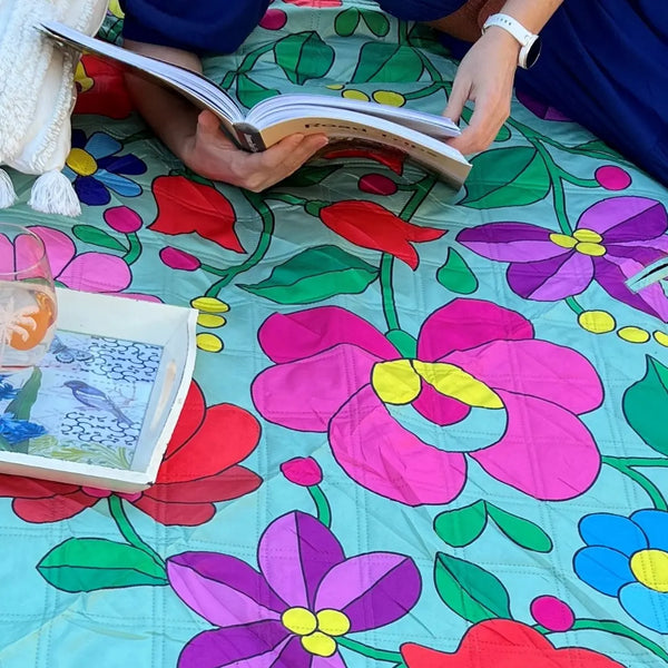 Close up of a woman lying on her side on a vibrant, flower-patterned picnic blanket, reading a book in a peaceful park setting. 