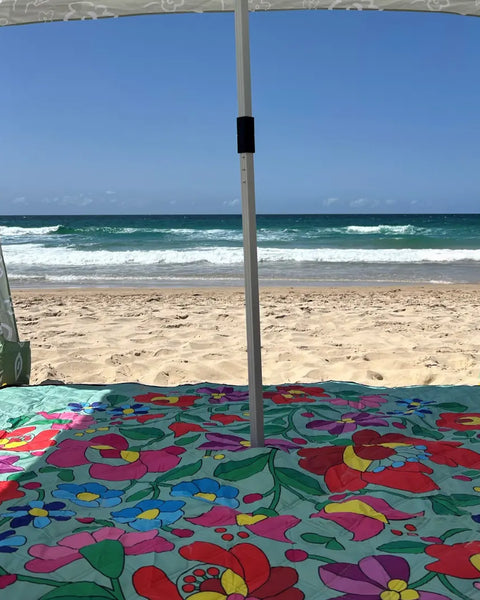 Nap and Sun Green-based picnic blanket with vivid flower pattern set up on the beach, with a sun cabana attached.