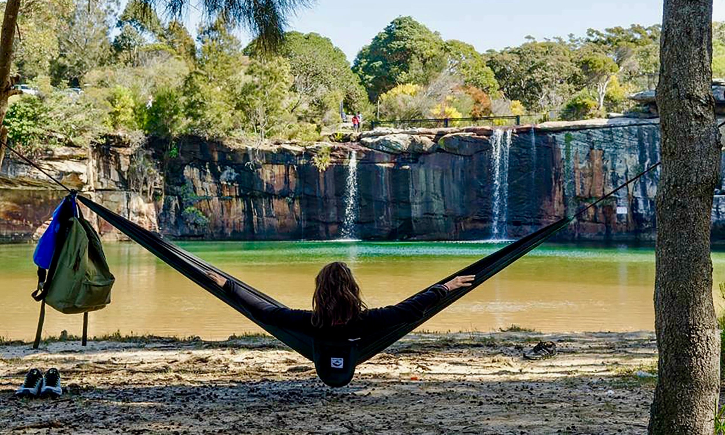 Person relaxing in a hammock by a scenic lake with a waterfall.