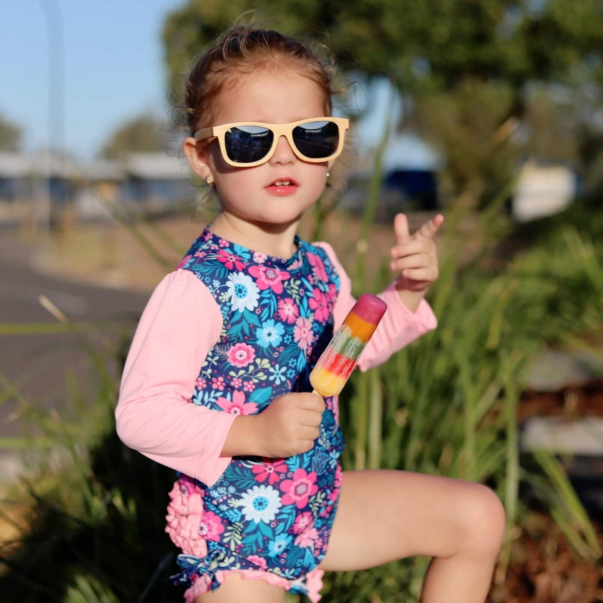 Child in a floral swimsuit and sunglasses holding a popsicle outdoors.