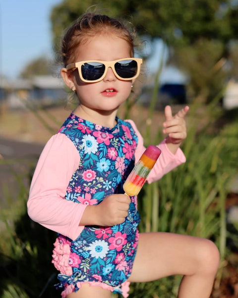 Child in a floral swimsuit and sunglasses holding a popsicle outdoors.