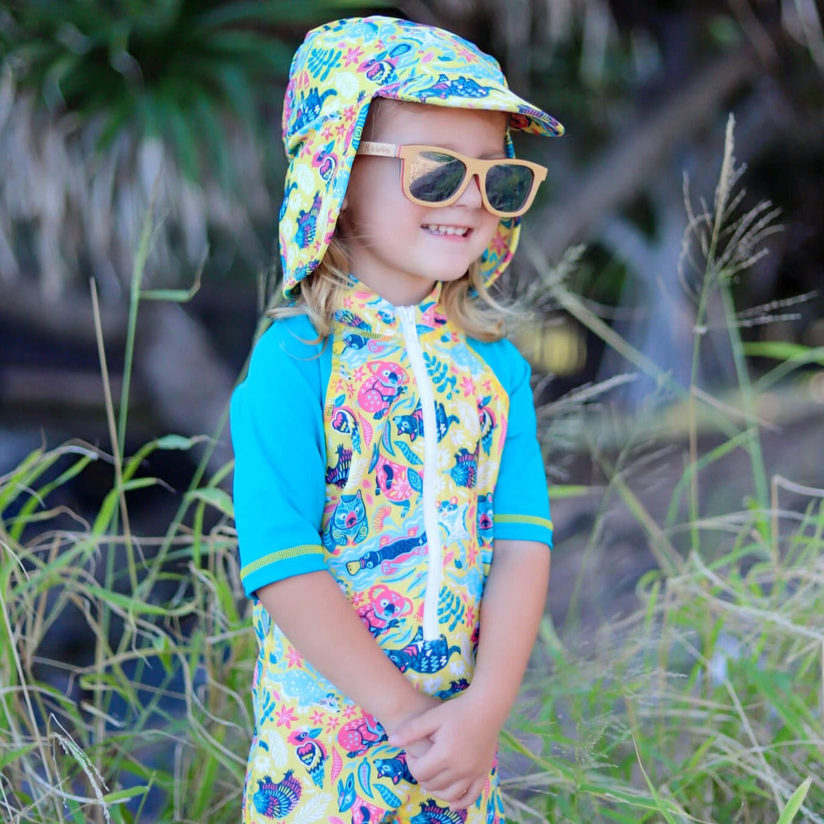 Child wearing a colorful swimsuit and hat with sunglasses, standing in a natural setting.
