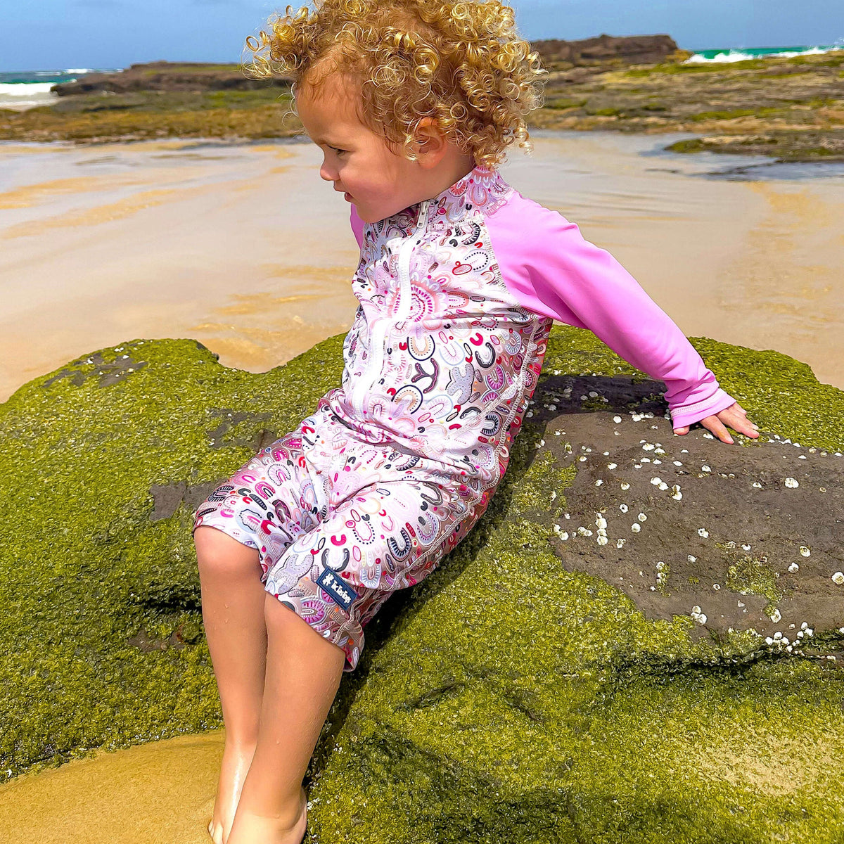 Child in a pink and white outfit sitting on a rock at the beach