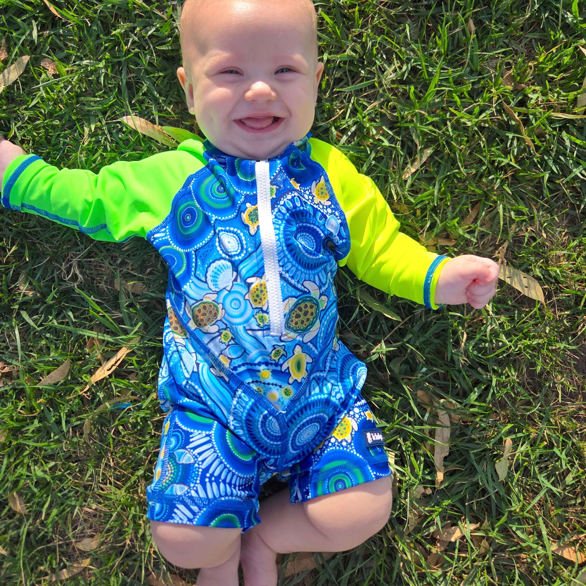 Baby wearing a blue and green swimsuit with turtle pattern on grass
