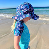 Child wearing a floral sun hat and blue shirt on a sandy beach with ocean in the background