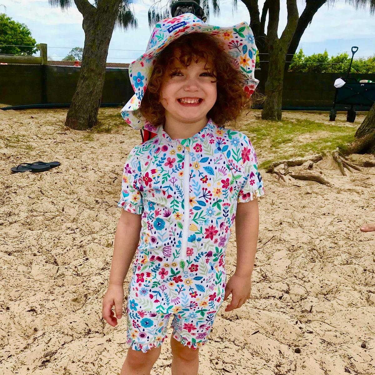 Child wearing a floral swimsuit and sun hat on a sandy beach.