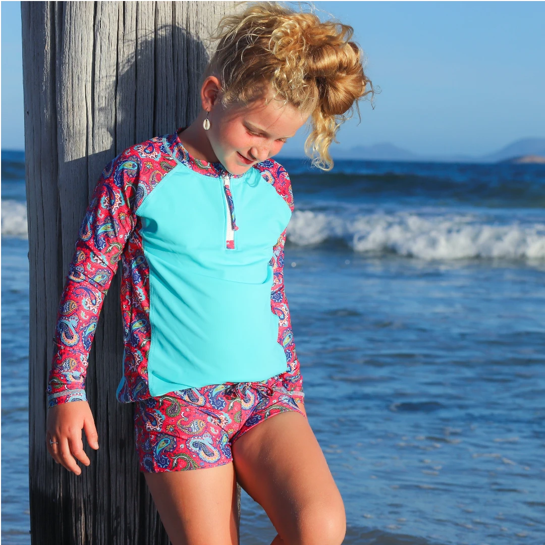 Young girl in a colorful swimsuit standing by a wooden post on a beach.
