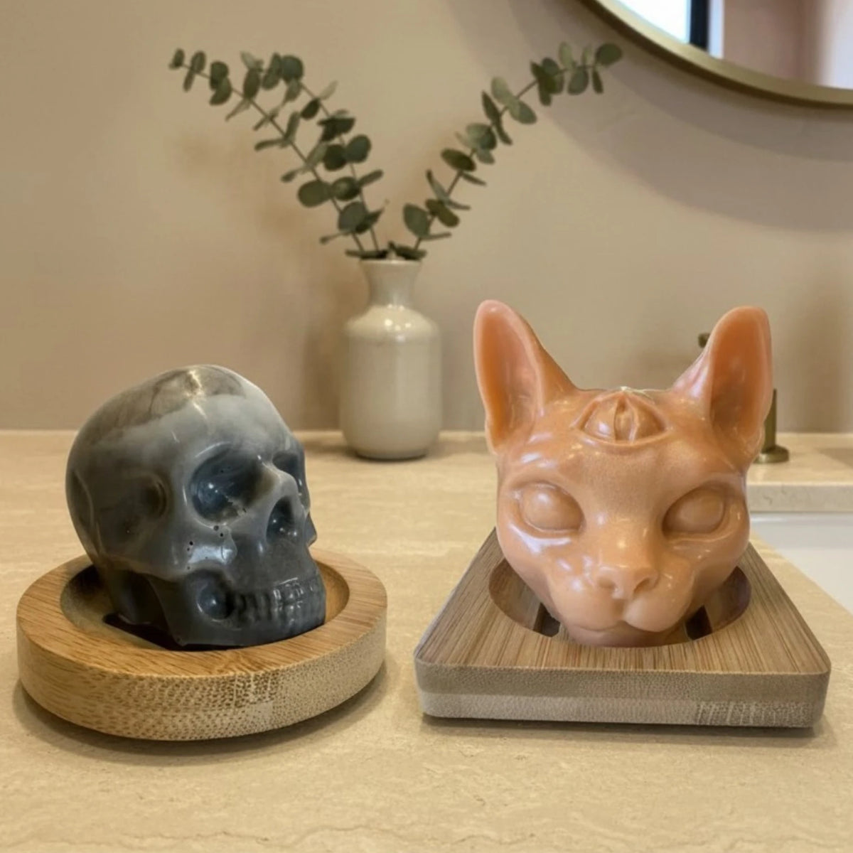 Two decorative head shaped objects displayed side by side on bamboo soap dishes in an indoor setting. The left object is a gray soap skull, the right object is a cat head shaped soap.