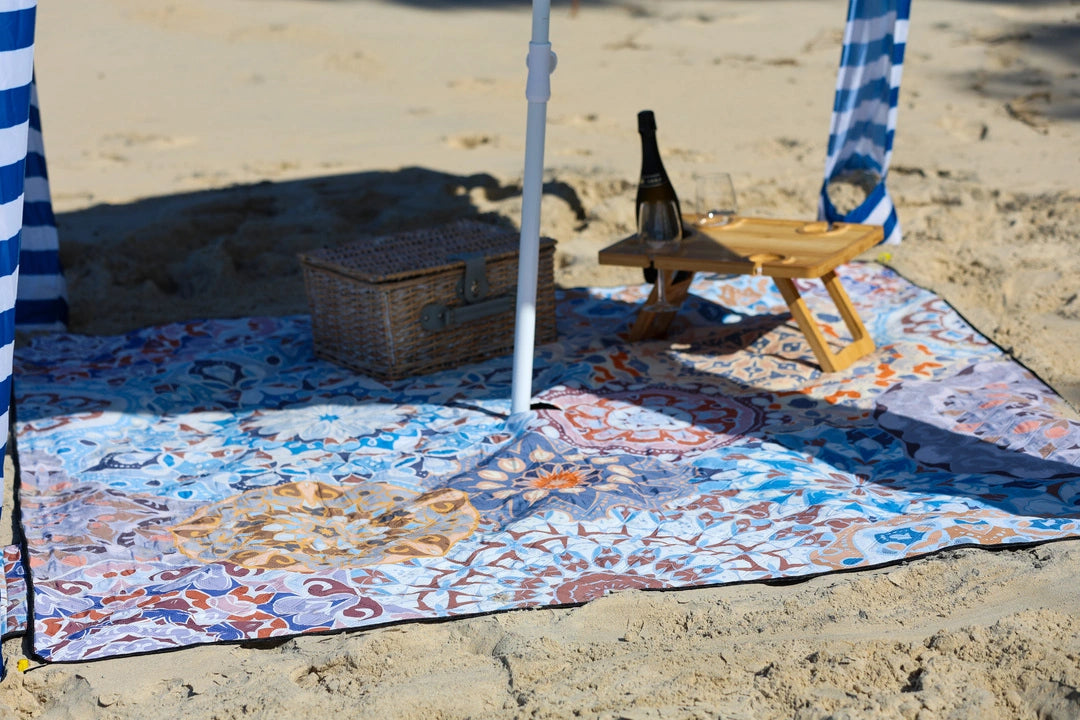 Beach setup with a mandala patterned blanket, picnic basket, and bottle on a sandy surface.