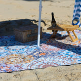 Beach setup with a mandala patterned blanket, picnic basket, and bottle on a sandy surface.