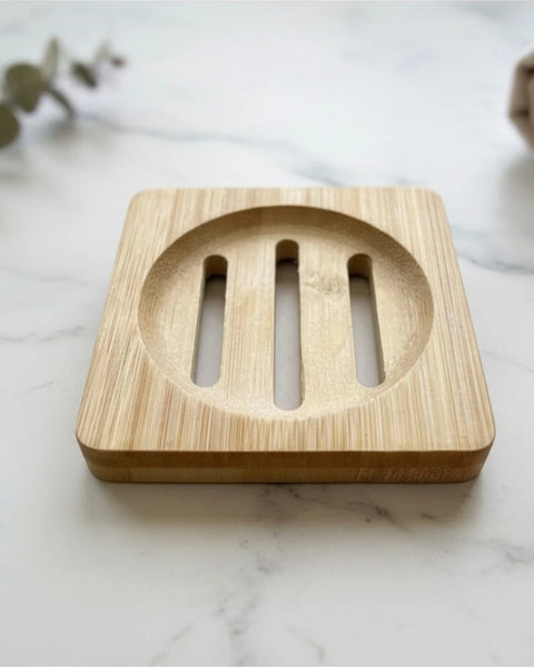A square bamboo soap dish with a circular center and three vertical slots, placed on a white marble surface with blurred greenery and a beige cloth in the background.