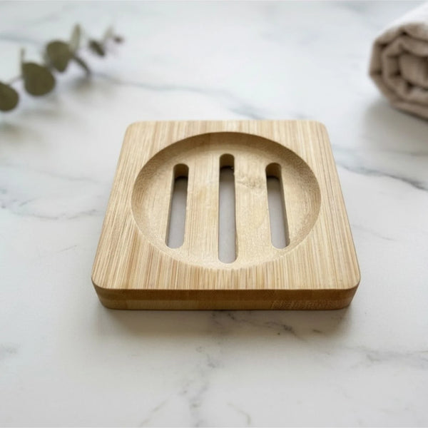 A square bamboo soap dish with a circular center and three vertical slots, placed on a white marble surface with blurred greenery and a beige cloth in the background.