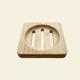 A square wooden soap dish with a circular recessed center and three vertical slits, shown on a plain light beige background.