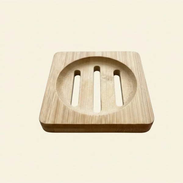 A square wooden soap dish with a circular recessed center and three vertical slits, shown on a plain light beige background.