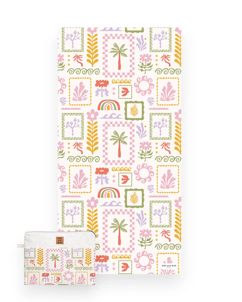 A view of a white beach towel featuring a colorful tropical patchwork pattern with palm trees, rainbows, flowers, and geometric frames and a matching bag.