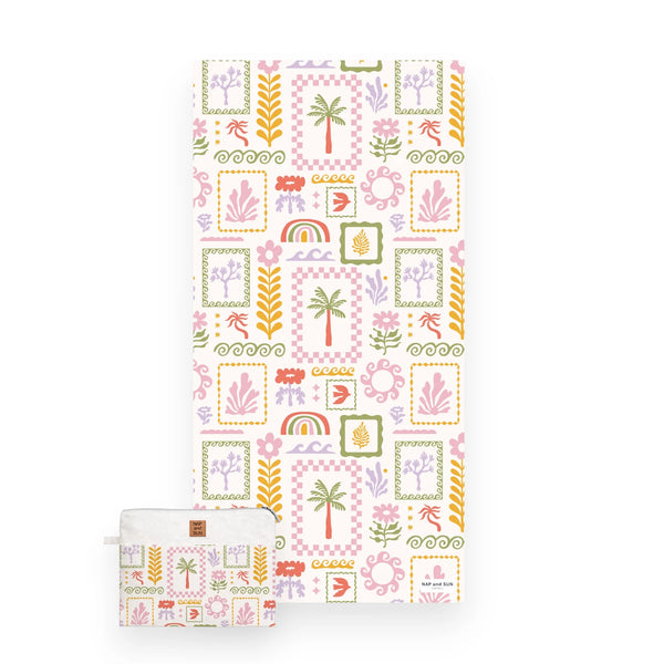 A view of a white beach towel featuring a colorful tropical patchwork pattern with palm trees, rainbows, flowers, and geometric frames and a matching bag.
