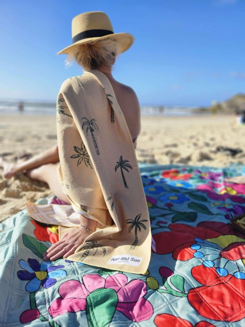 Woman seen from behind sitting on a floral Nap and Sun brand waterproof picnic blanket at the beach, with a palm-pattern sand-free beach towel draped over her shoulder. 