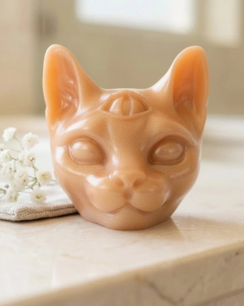A sculpted cat head soap in a warm peach-beige tone, photographed on a light stone surface with small white flowers beside it in a softly blurred indoor setting.