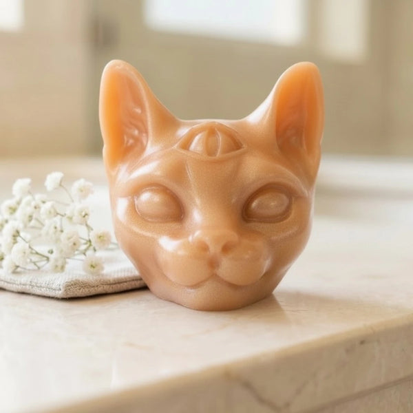 A sculpted cat head soap in a warm peach-beige tone, photographed on a light stone surface with small white flowers beside it in a softly blurred indoor setting.