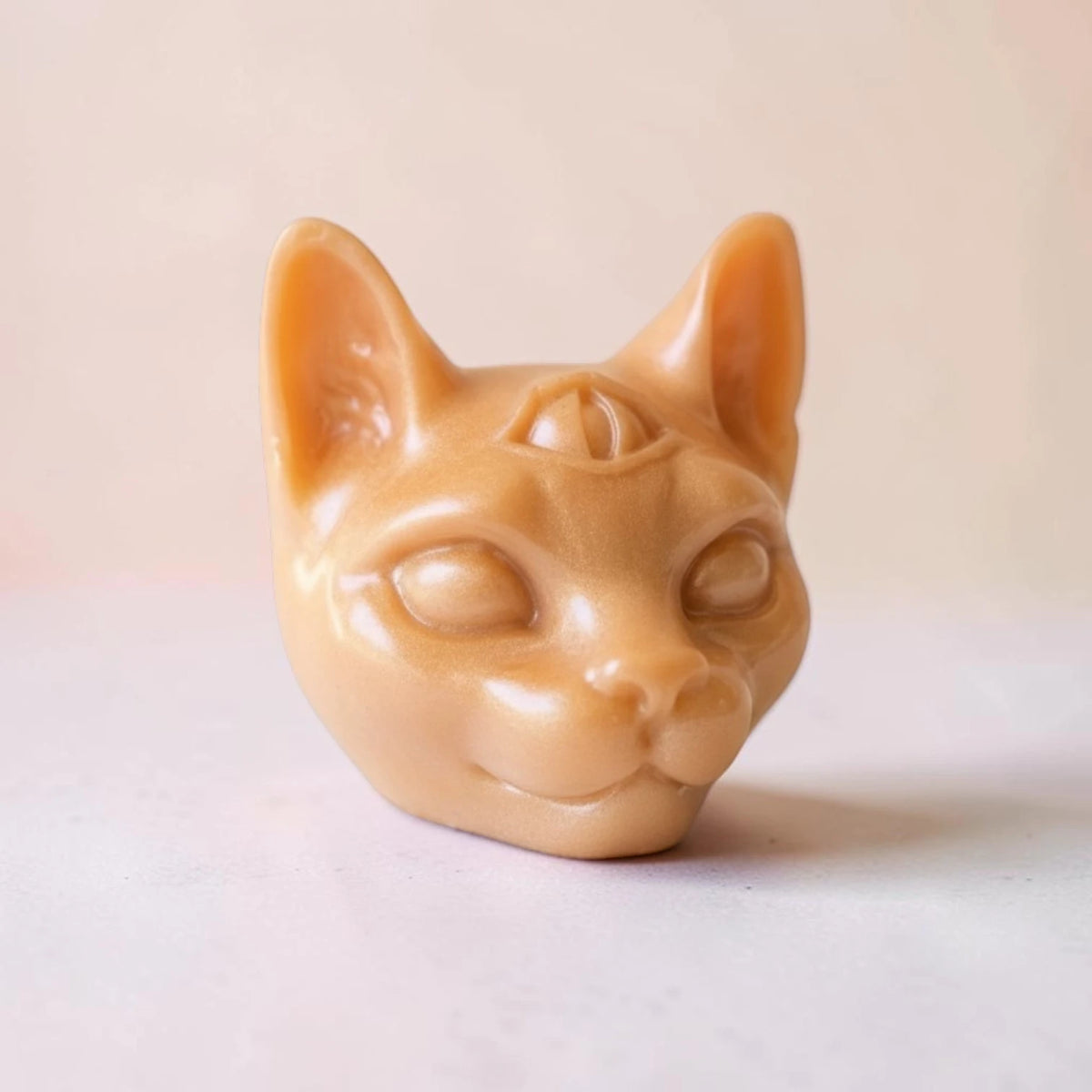 A small sculpted cat head soap in a warm tan or peachy-beige tone, photographed against a soft light beige background.