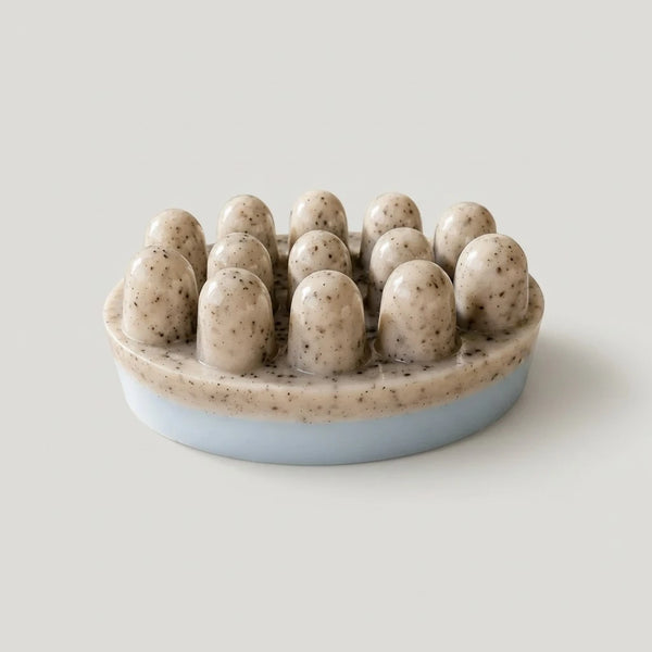 An oval shaped massage soap bar with a light blue base and beige, photographed on a neutral light gray background.