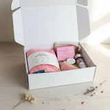 Open white box containing pink hair wrap towel and other bath pamper items on a light surface.