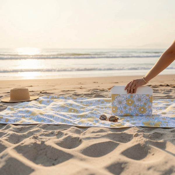 A beach scene at sunset with a beach towel featuring a yellow and white daisy pattern spread on the sand. A person's arm is placing a matching patterned bag, with a straw hat and sunglasses nearby.
