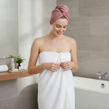 Woman in a bathroom with a pink towel on her head and a white towel wrapped around her, smiling.