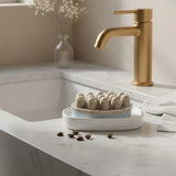 A matte gold single-handle bathroom faucet mounted on a white marble countertop next to a white sink, with a small ceramic dish holding massage soap bar and a blurred vase of flowers in the background.