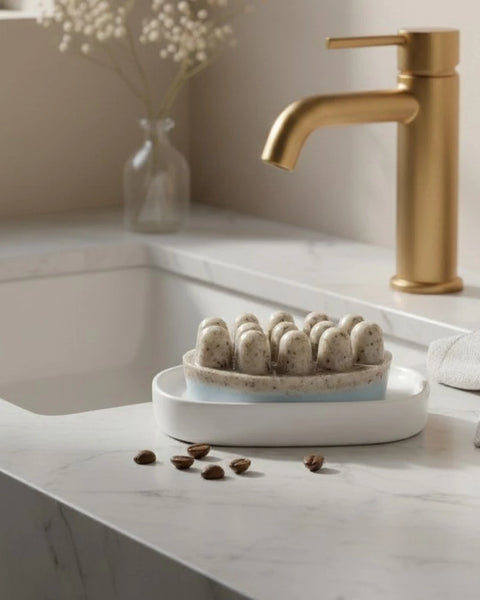A matte gold single-handle bathroom faucet mounted on a white marble countertop next to a white sink, with a small ceramic dish holding massage soap bar and a blurred vase of flowers in the background.