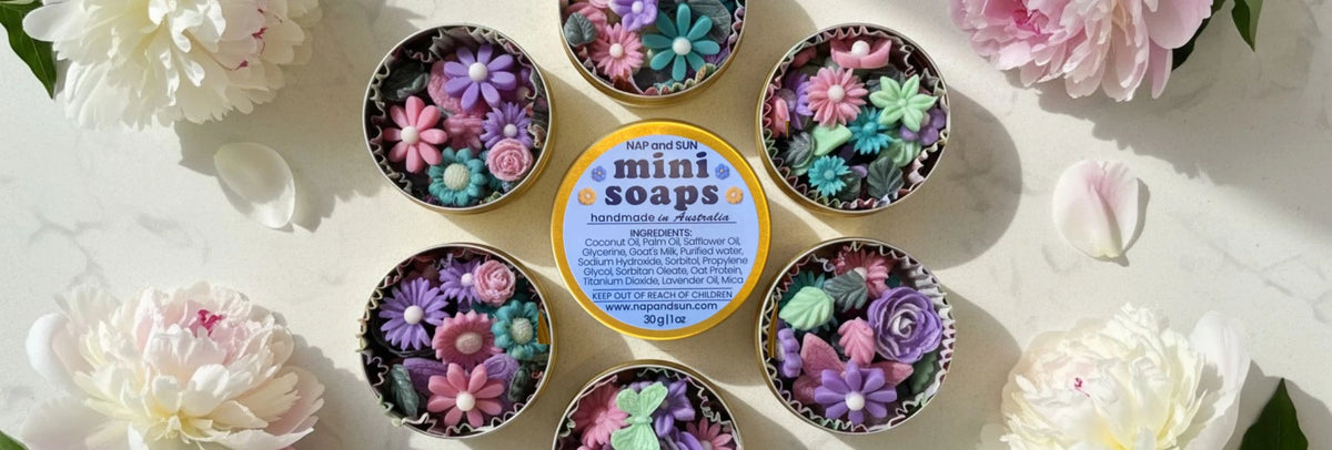  Golden round tins filled with colorful floral-shaped mini soaps, arranged on white marble and surrounded by pastel peonies. The centre tin features “MINI SOAPS” on the lid.